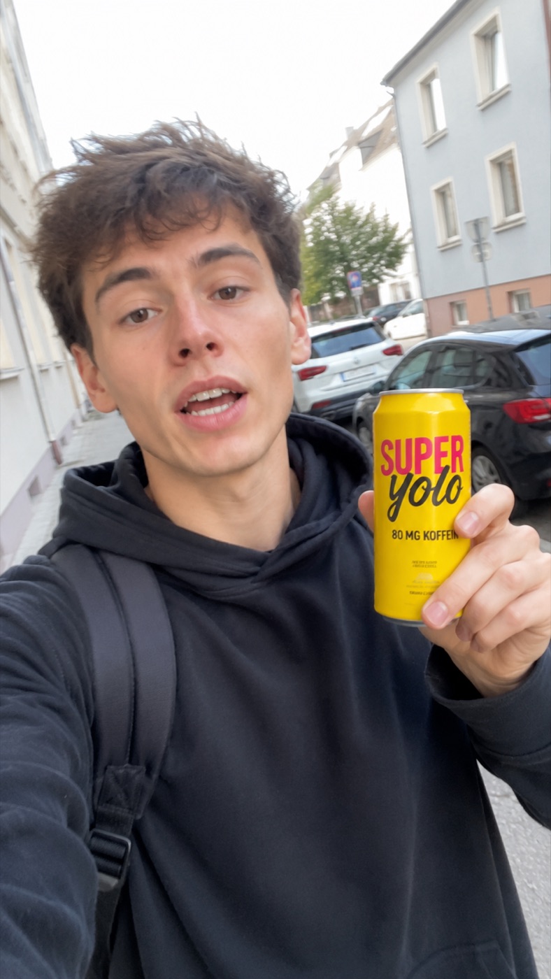 Junger Mann hält auf der Straße eine gelbe SUPERYOLO-Dose in die Handykamera