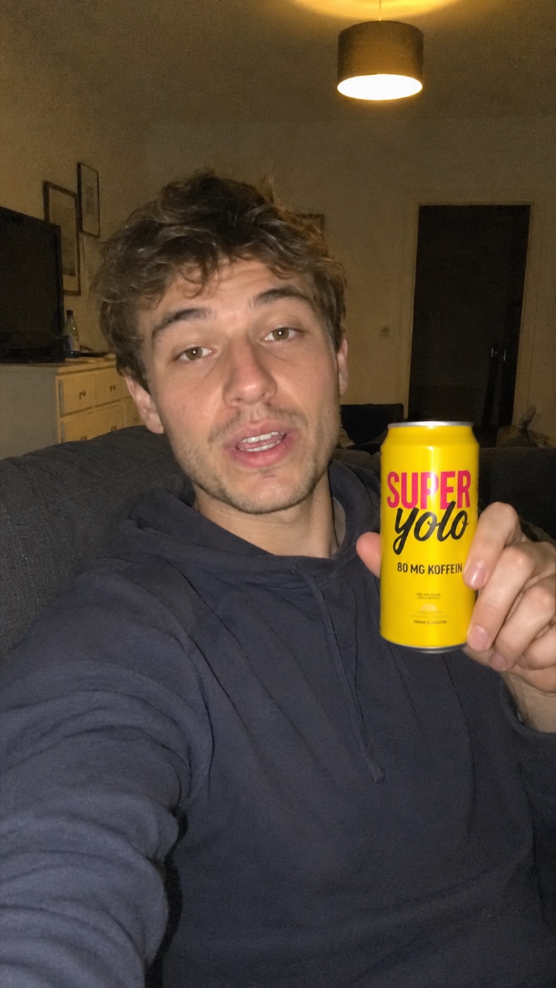 Junger Mann hält abends im Wohnzimmer eine gelbe SUPERYOLO-Dose in die Kamera